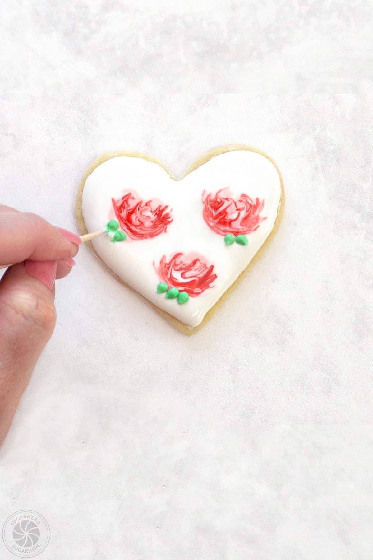 Hand using a toothpick to extend light green dots of icing to look like rose leaves.
