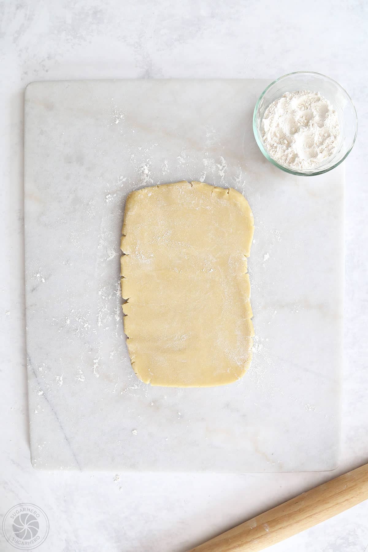 A disc of sugar cookie dough rolled into a rectangle on a marble cutting board next to flour and a rolling pin.