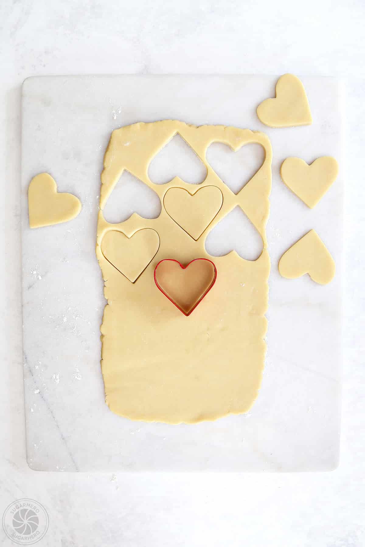 Sugar cookie dough being cut into hearts with a red heart shaped cookie cutter.