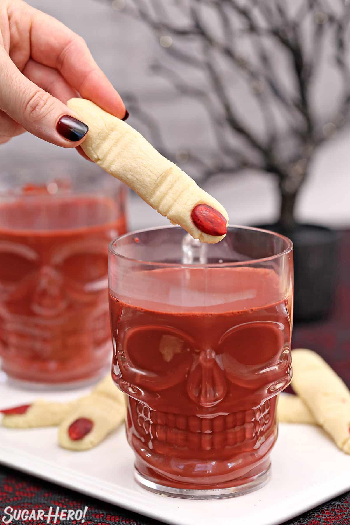 Hand holding a finger-shaped cookie about to dunk it in a mug of red velvet hot chocolate.