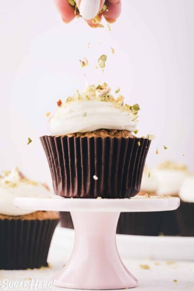 Hand sprinkling crushed pistachios on top of a zucchini cupcake frosted with cream cheese frosting.